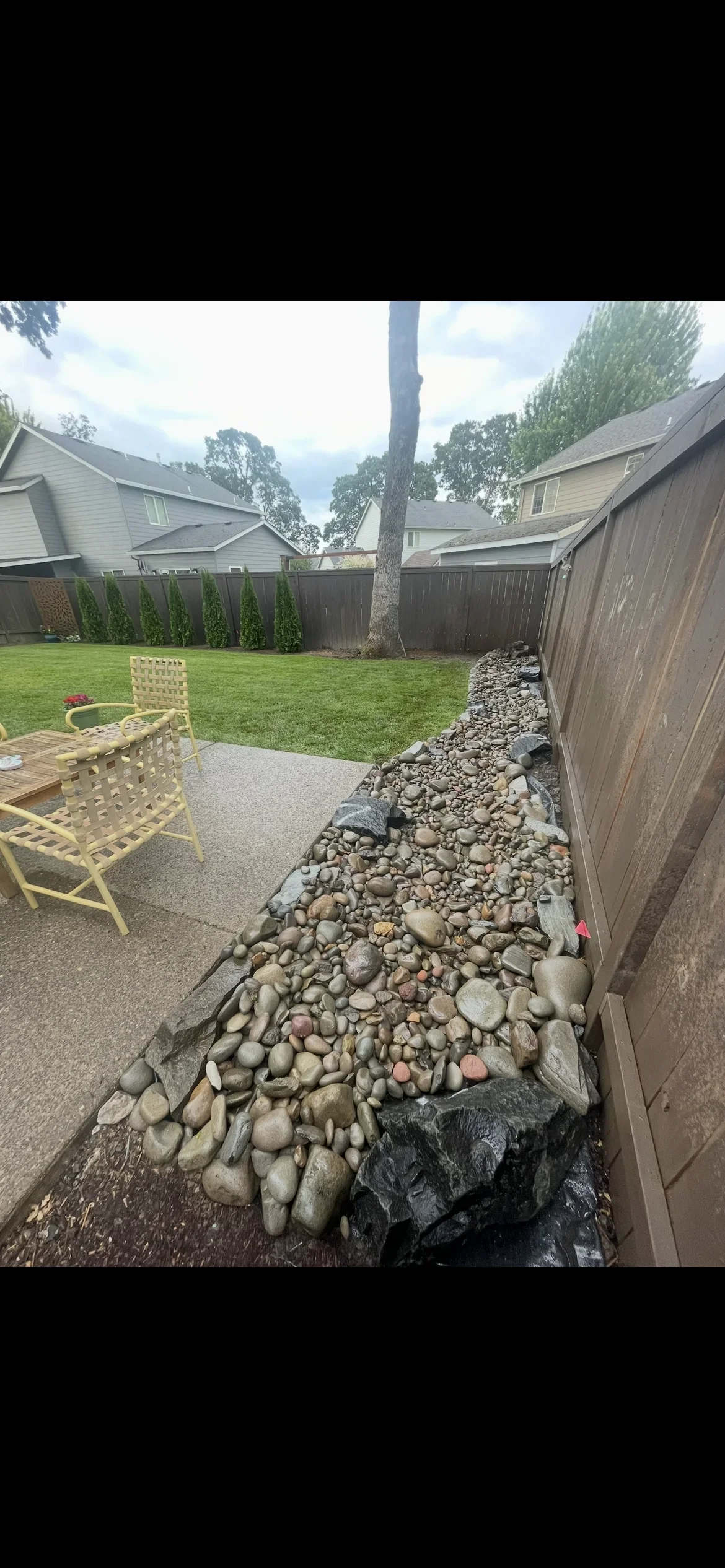River rock drainage bed with landscape fabric along fence, concrete patio, and manicured lawn