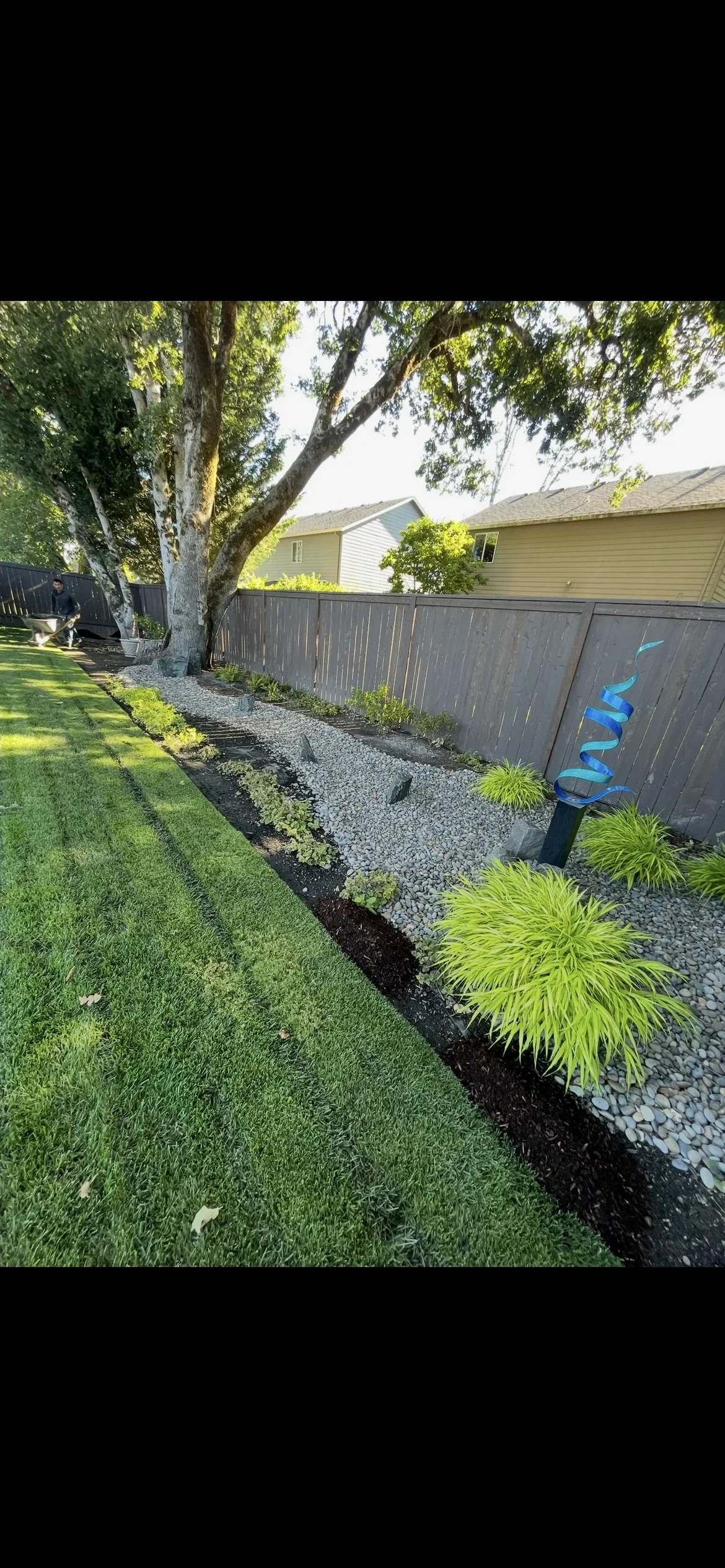 Landscape bed with river rock, black mulch border, and ornamental grasses along fence line